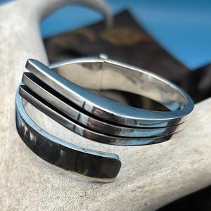 May include: A silver-toned cuff bracelet with a modern design. The bracelet features three parallel bands and an open, wrap-around style. The polished metal reflects light, creating a sleek and contemporary look. Jewelry.