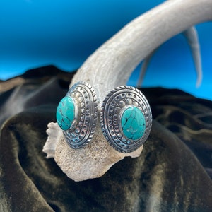 May include: A pair of silver earrings with turquoise stones set in a circular design. The earrings have a textured, ornate border.