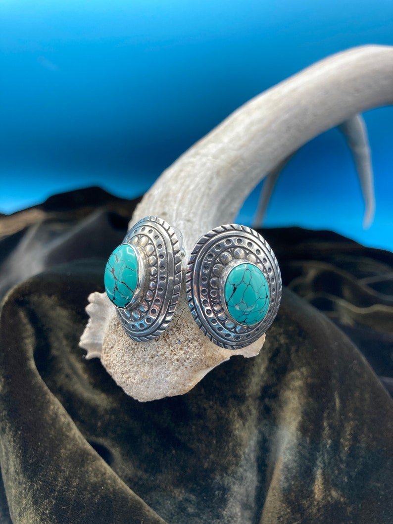 May include: A pair of silver earrings with turquoise stones set in a circular design. The earrings have a textured, ornate border.