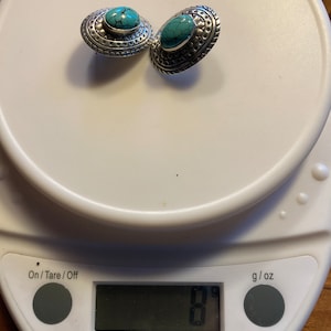 May include: A pair of silver earrings with turquoise stones set in a decorative design. The earrings are on a digital scale that reads 8 grams.