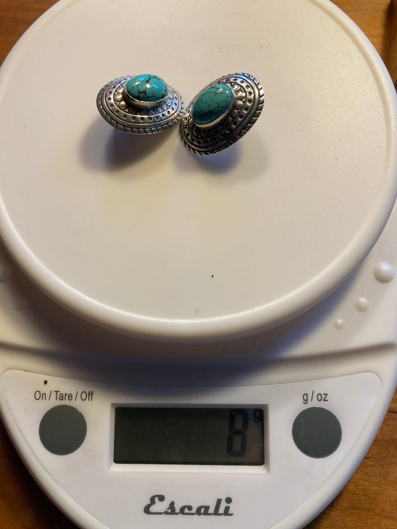May include: A pair of silver earrings with turquoise stones set in a decorative design. The earrings are on a digital scale that reads 8 grams.