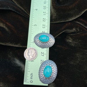 May include: Two oval-shaped turquoise and silver earrings with a decorative border. The earrings are approximately 2.5 cm in length.