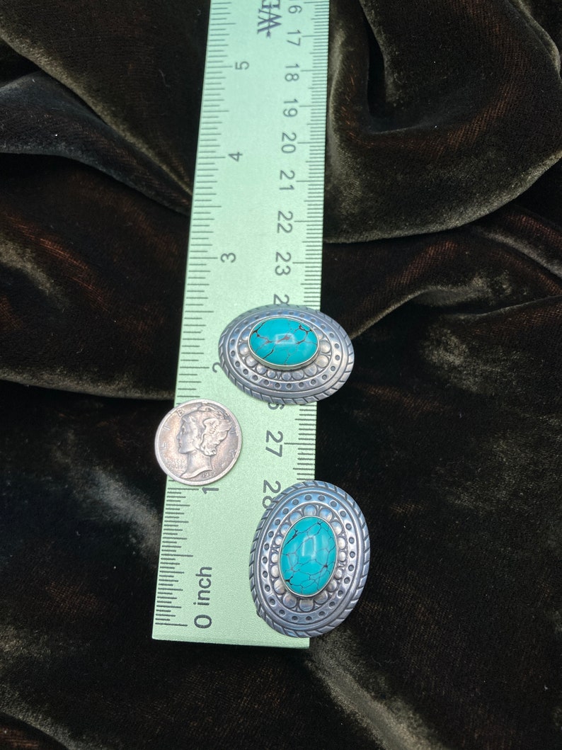 May include: Two oval-shaped turquoise and silver earrings with a decorative border. The earrings are approximately 2.5 cm in length.