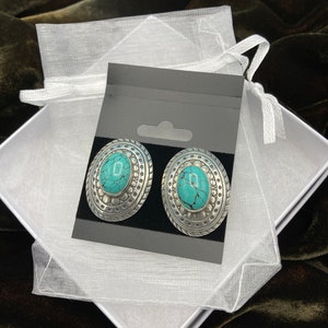 May include: A pair of silver earrings with turquoise stones set in a decorative oval design. The earrings are displayed on a black card with a white background.