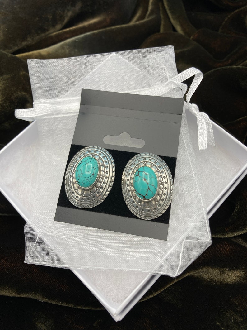 May include: A pair of silver earrings with turquoise stones set in a decorative oval design. The earrings are displayed on a black card with a white background.