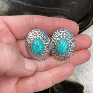 May include: A pair of silver oval earrings with turquoise stones set in the centre. The earrings have a detailed design around the stones.