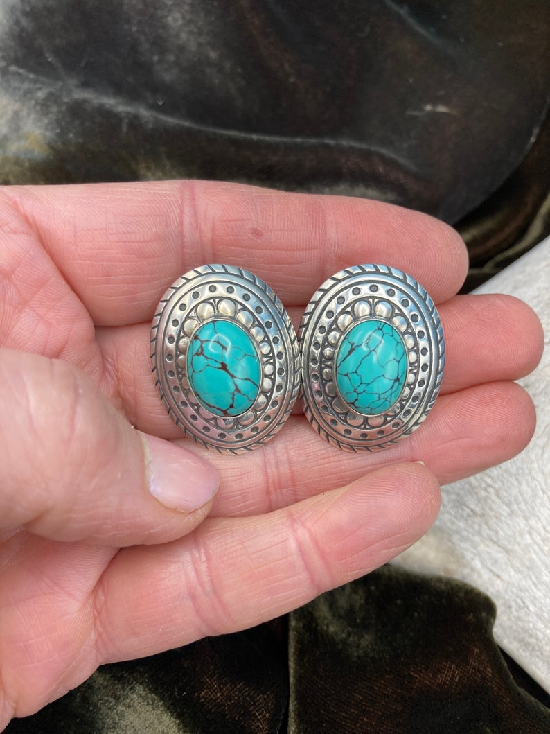 May include: A pair of silver oval earrings with turquoise stones set in the centre. The earrings have a detailed design around the stones.