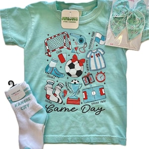 May include: A light blue t-shirt with a soccer-themed graphic, including a soccer ball, cleats, and a soccer field. The shirt is paired with white and light blue socks, and a matching hair bow. The text "Game Day" is printed on the shirt.