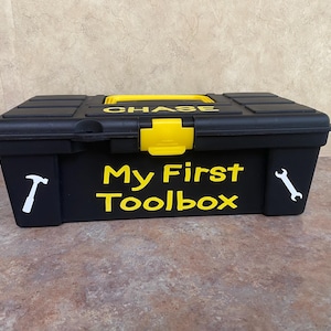 May include: Black plastic toolbox with yellow accents and the words "My First Toolbox" in yellow lettering. The toolbox has a white hammer and wrench on the sides and the name "CHASE" on the top.
