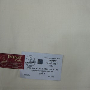 May include: A white fabric swatch with a red and gold label that reads "Vaishali Lohi" and a smaller label that reads "Dharyawal". The larger label also includes the text "274 x 137 cm" and "Dharyawal". The smaller label includes the text "Dharyawal" and a logo of a rooster. Below the labels is a white card with text in Hindi that reads "Dharyawal "Vaishali Lohi" Lohi" and "Lambai 274 cm. X Chaudaai 137 cm. Drycleaning upayukt pakka rang tukde! Bharat me bana"