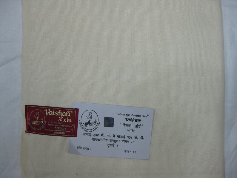 May include: A white fabric swatch with a red and gold label that reads "Vaishali Lohi" and a smaller label that reads "Dharyawal". The larger label also includes the text "274 x 137 cm" and "Dharyawal". The smaller label includes the text "Dharyawal" and a logo of a rooster. Below the labels is a white card with text in Hindi that reads "Dharyawal "Vaishali Lohi" Lohi" and "Lambai 274 cm. X Chaudaai 137 cm. Drycleaning upayukt pakka rang tukde! Bharat me bana"