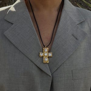 May include: A gold cross pendant necklace with clear crystal accents on a black cord. The necklace is worn by a person wearing a gray blazer.