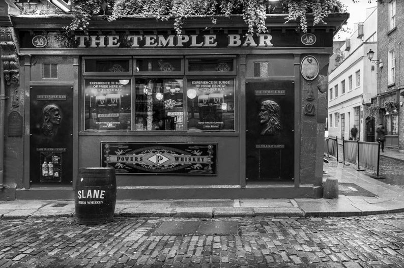Puede incluir: Una foto en blanco y negro del pub The Temple Bar en Dubl&iacute;n, Irlanda. El pub tiene un letrero sobre la entrada que dice "The Temple Bar". Hay un letrero en el lateral del edificio que dice "The Temple Bar" y presenta un retrato de un hombre. Hay un barril frente al pub que dice "Slane Irish Whiskey".