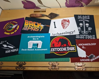 May include: A collection of eight colorful iron-on patches with various designs and text related to archaeology. The patches feature a desk, a mammoth, a pickaxe, a heart, and a rainbow. The text on the patches includes "Back to the Past", "Archaeologists Do It", "Black Mattock", "I Love Archaeology", "Leistocene Park", "Archaeology is... Ground Breaking", and "Through Time and Space".