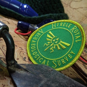 May include: A green and yellow embroidered patch with the text "Hyrule Royal Archaeological Survey" and a stylised Triforce symbol.