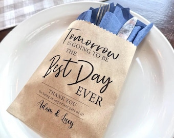 May include: A brown paper pouch containing a blue napkin, a metal fork, and a metal knife. The pouch features black text that reads: Tomorrow is going to be the best day ever. Thank you for being an important part of it! Adam & Lucy.