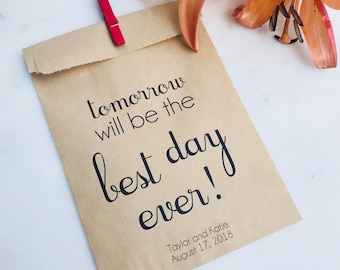 May include: A brown paper bag with the text "tomorrow will be the best day ever!" in black script. A red clothespin secures the bag's top. Peach-colored lilies with brown spots are in the background. The date "August 17, 2018" is printed on the bag.