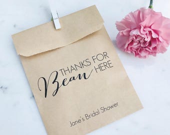 May include: A small, tan paper bag with the text "THANKS FOR BEAN HERE" and "Jane's Bridal Shower" printed in black. A white clothespin secures the bag's flap. A pink carnation flower is to the right.