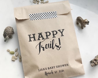 May include: A brown paper bag with the text "HAPPY Trails!" in black, sealed with a black and white polka dot sticker. The bag is labeled "LISA'S BABY SHOWER March 17. 2018". Decorative animal figurines and seeds are scattered around the bag.