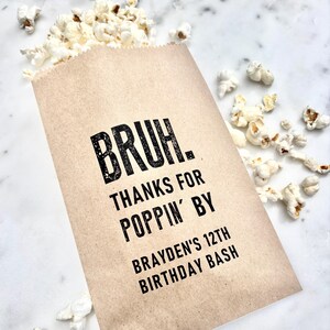 May include: A brown paper popcorn bag with the text "BRUH. THANKS FOR POPPIN' BY BRAYDEN'S 12TH BIRTHDAY BASH" in black. The bag is filled with white popcorn, with some kernels scattered around on a white marble surface.