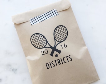 May include: A brown paper bag sealed with a strip of black and white polka dot tape. The bag features a black graphic of two crossed tennis rackets, the number 2016, and the word "DISTRICTS".