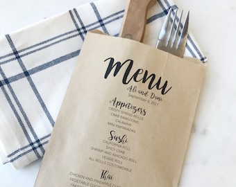 May include: A kraft paper menu bag with the words "Menu" and "Ali and Dani" printed in black script. The menu lists appetizers, sushi, Thai dishes, and beverages. A wooden knife and fork rest inside the bag, which is placed on a blue and white checkered napkin.