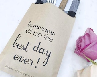 May include: A brown paper bag with the text "tomorrow will be the best day ever!" and "Sanya & Ryan's Rehearsal Dinner" printed in black. Inside the bag are a fork, a knife, and a black and white checkered napkin. A pink rose and a cream rose are in the background.