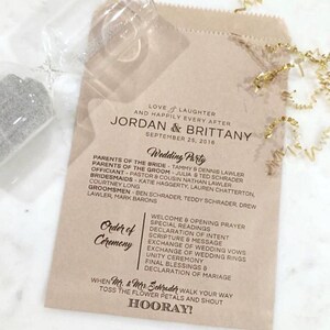 Puede incluir: Un programa de boda impreso en una bolsa de papel marrón. El programa enumera los nombres de los novios, la fiesta de bodas y el orden de la ceremonia. Cinta dorada y un vaso transparente en el fondo.