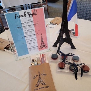 May include: A table setting for a French-themed dinner party. A brown paper bag with a printed Eiffel Tower and the text "Bon Jour! Cafe de Heritage" is on the table. There are also black and white macarons in clear plastic containers with black and white ribbon bows.