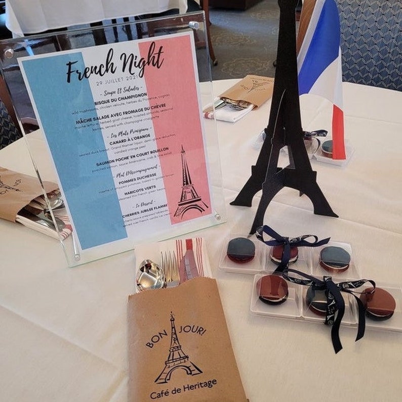 May include: A table setting for a French-themed dinner party. A brown paper bag with a printed Eiffel Tower and the text "Bon Jour! Cafe de Heritage" is on the table. There are also black and white macarons in clear plastic containers with black and white ribbon bows.