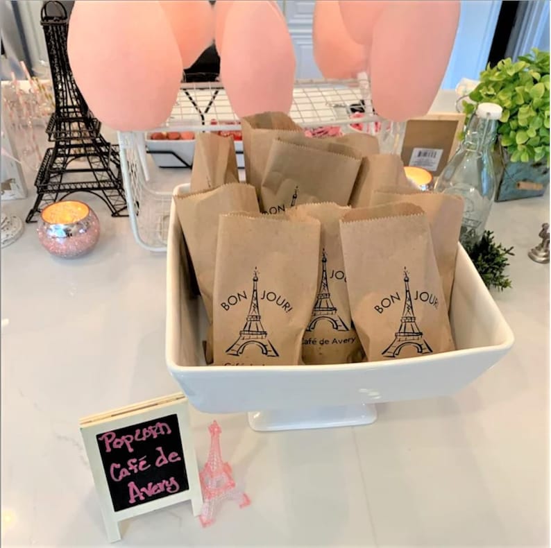 May include: A white ceramic bowl filled with brown paper bags. Each bag has a black and white Eiffel Tower design with the text "Bon Jour" printed on it. A small black chalkboard sign reads "Popcorn Cafe de Avery".