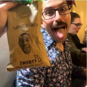 Puede incluir: Una bolsa de papel marr&oacute;n con el texto "Look Who's Thirty" y una cara de dibujos animados impresa en ella. La bolsa est&aacute; siendo sostenida por una persona que lleva una camisa floral y gafas.