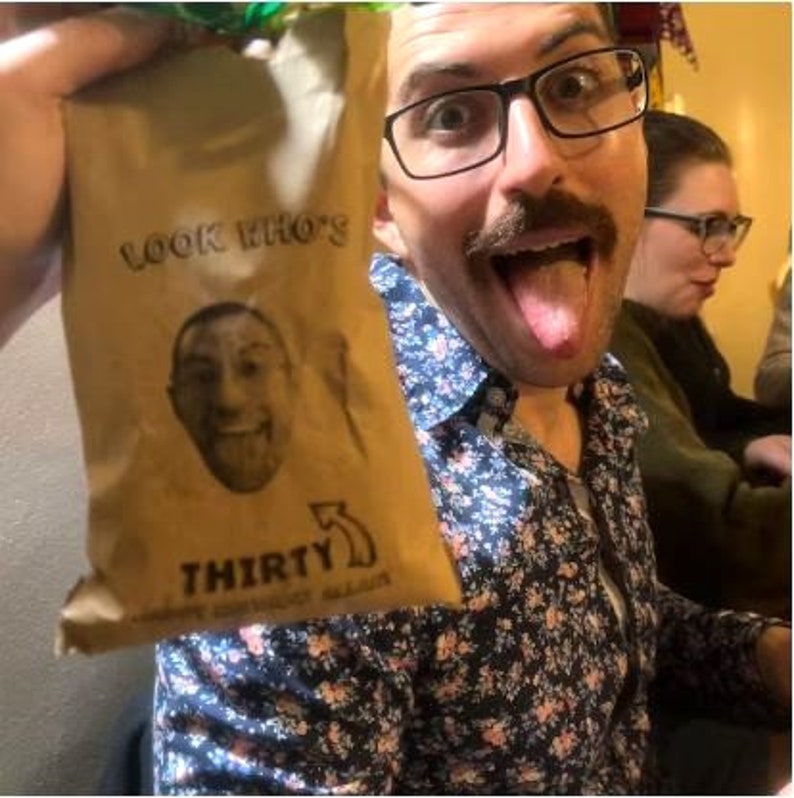 Puede incluir: Una bolsa de papel marr&oacute;n con el texto "Look Who's Thirty" y una cara de dibujos animados impresa en ella. La bolsa est&aacute; siendo sostenida por una persona que lleva una camisa floral y gafas.