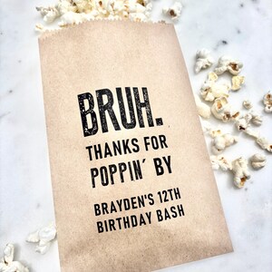 May include: A brown paper popcorn bag with the text "BRUH. THANKS FOR POPPIN' BY BRAYDEN'S 12TH BIRTHDAY BASH" in black. The bag is filled with popcorn, with some kernels scattered around.