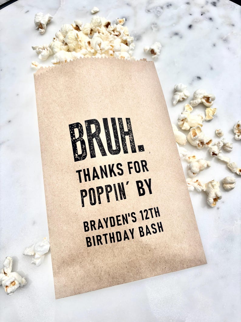 May include: A brown paper popcorn bag with the text "BRUH. THANKS FOR POPPIN' BY BRAYDEN'S 12TH BIRTHDAY BASH" in black. The bag is filled with popcorn, with some kernels scattered around.