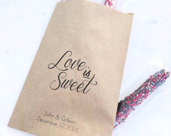 May include: A brown paper bag with the text "Love is Sweet" in elegant script. The bag contains chocolate-covered pretzel rods with red and white sprinkles. The bag also includes the names "John & Colleen" and the date "December 12, 2025".
