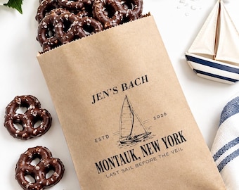 May include: A brown paper bag filled with chocolate-covered pretzels, printed with a sailboat graphic and the text: JEN'S BACH, ESTD 2026, MONTAUK, NEW YORK, LAST SAIL BEFORE THE VEIL. The bag is surrounded by loose chocolate pretzels and chunks.