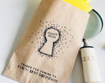 May include: A brown paper bag with the words "ESCAPE ROOM" inside a keyhole design. The bag also says "THANKS FOR COMING TO EVIE'S 11TH BIRTHDAY!" Yellow tissue paper and blue game pieces peek out from the top. A magnifying glass is on the right.