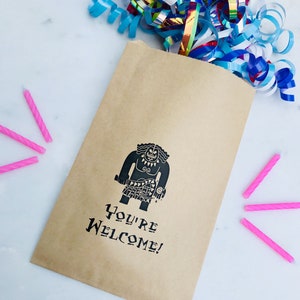 May include: A brown paper bag with a black and white illustration of Maui from the movie Moana. The bag has the text "You're Welcome!" printed on it. There are pink birthday candles on a white surface next to the bag.