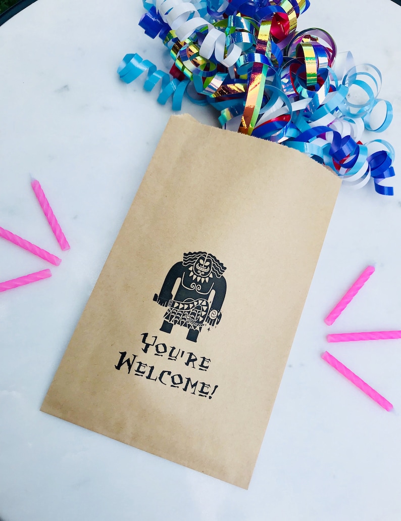 May include: A brown paper bag with a black and white illustration of Maui from the movie Moana. The bag has the text "You're Welcome!" printed on it. There are pink birthday candles on a white surface next to the bag.