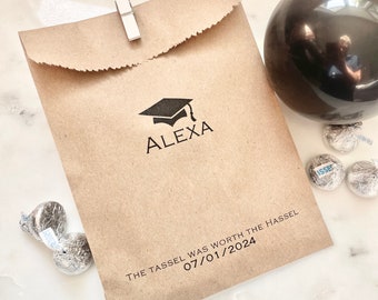 May include: A brown paper bag secured with a white clip, featuring a black graduation cap icon, the name ALEXA, and the text THE TASSEL WAS WORTH THE HASSEL 07/01/2024. A black balloon and several silver-wrapped chocolate candies are placed nearby.