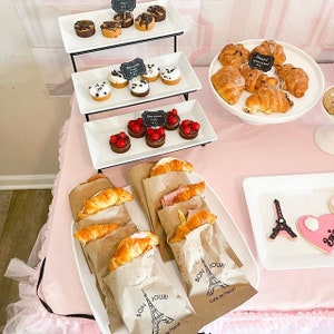 May include: A pink table setting with a variety of pastries, including croissants, mini cupcakes, and pastries with red fruit toppings. The table is decorated with a Parisian theme, including a miniature Eiffel Tower and the phrase "Bon Jour!" on the brown paper bags.