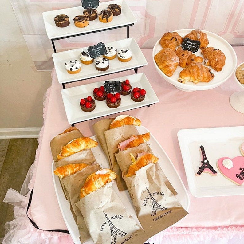 May include: A pink table setting with a variety of pastries, including croissants, mini cupcakes, and pastries with red fruit toppings. The table is decorated with a Parisian theme, including a miniature Eiffel Tower and the phrase "Bon Jour!" on the brown paper bags.
