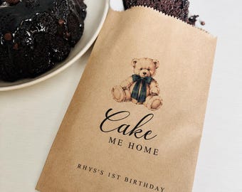 May include: A light brown paper bag featuring a printed illustration of a teddy bear with a green plaid bow. The text reads Cake ME HOME and RHYS'S 1ST BIRTHDAY. A slice of dark chocolate cake with chocolate chips sits inside the bag.