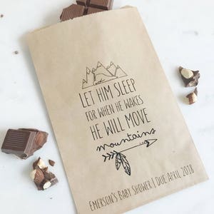 May include: A brown paper gift bag with the text "LET HIM SLEEP FOR WHEN HE WAKES HE WILL MOVE mountains". The bag has a mountain and arrow design. Chocolate pieces are scattered around the bag. The text "EMERSON'S BABY SHOWER | DUE APRIL 2018" is at the bottom.
