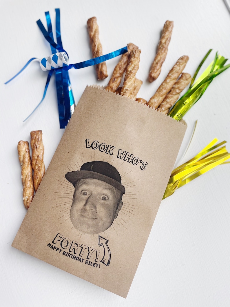 May include: A brown paper bag with a birthday message printed on it. The message reads 'Look Who's Forty! Happy Birthday Riley!' and features a black and white photo of a person wearing a hat. The bag is filled with pretzel sticks and has blue and yellow streamers coming out of the top.