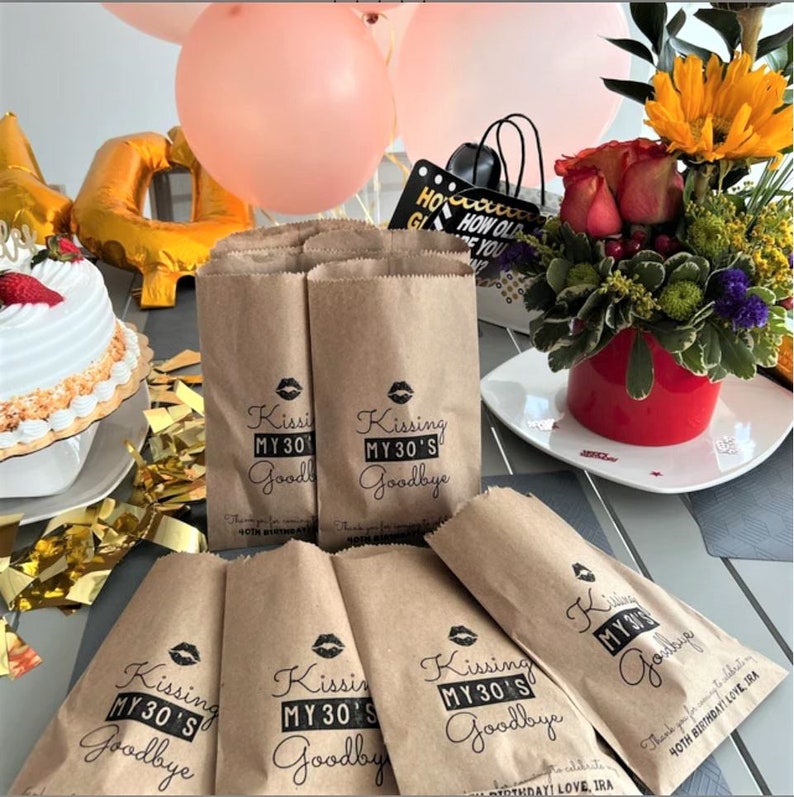 Puede incluir: Bolsas de papel marr&oacute;n con texto negro y una impresi&oacute;n de labios rojos que dice "Kissing My 30's Goodbye". Las bolsas est&aacute;n sobre una mesa con un pastel, globos y flores.