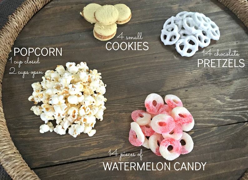 Puede incluir: Una bandeja de madera con una variedad de aperitivos, incluyendo palomitas de ma&iacute;z, galletas, pretzels y caramelos de sand&iacute;a. La bandeja est&aacute; decorada con un borde de cuerda trenzada. El texto en la bandeja dice: "POPCORN 1 cup closed 2 cups open", "4 small COOKIES", "14 chocolate PRETZELS", y "14 pieces of WATERMELON CANDY".