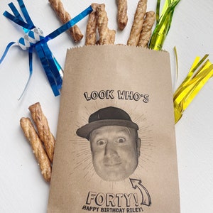 Puede incluir: Una bolsa de papel marr&oacute;n con el texto "Look Who's Forty! Happy Birthday Riley!" impreso en ella. Un rostro de hombre est&aacute; impreso en la bolsa, y lleva una gorra de b&eacute;isbol. Hay palitos de pretzel y serpentinas azules y amarillas en la mesa.
