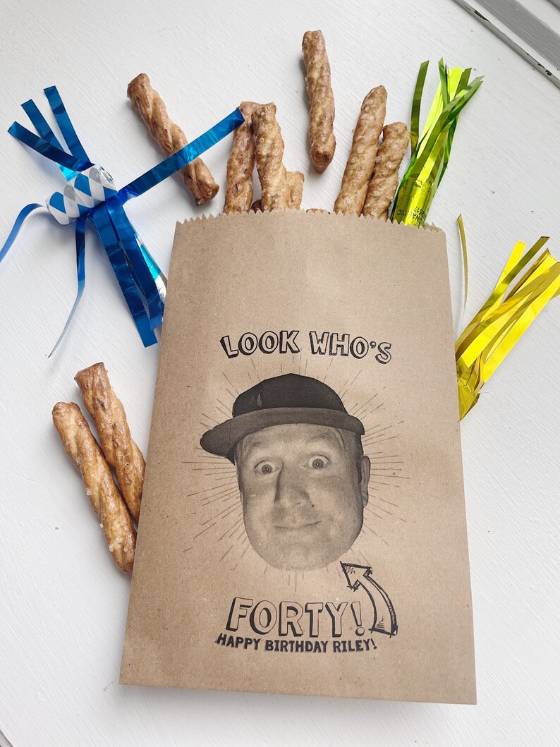 Puede incluir: Una bolsa de papel marr&oacute;n con el texto "Look Who's Forty! Happy Birthday Riley!" impreso en ella. Un rostro de hombre est&aacute; impreso en la bolsa, y lleva una gorra de b&eacute;isbol. Hay palitos de pretzel y serpentinas azules y amarillas en la mesa.